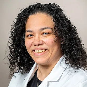 Jennifer Abdullah has long, dark curly hair, is wearing a black blouse under a white medical coat, and is smiling at the camera.