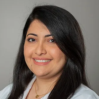 Dr. Al Allawi has long dark straight hair, is wearing a necklace and dark blouse under a white medical coat, and is smiling at the camera.
