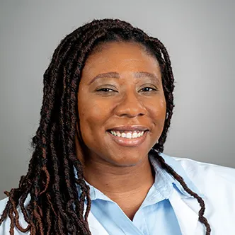 LaToya Augustine has long dark hair styled in long braids, is wearing a light blue blouse under a white medical coat, and is smiling at the camera.
