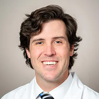 Dr. Bentz has short wavy brown hair, is wearing a white dress shirt and blue striped tie under a white medical coat, and is smiling at the camera.