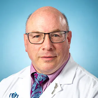 Dr. Berney is wearing glasses and a purple shirt with blue patterned tie under a white medical coat. He is looking at the camera.