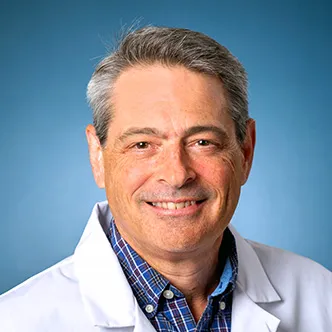 Dr. Britton has short gray-black hair, is wearing a white medical coat and blueish shirt, and is smiling at the camera.