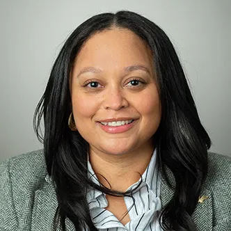Tessa Burrus-Sanders has long wavy dark hair, is wearing a gray suit jacket and blue blouse, and is smiling at the camera.