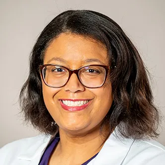 Amber Copeland has wavy dark brown hair, is wearing glasses and a purple blouse under a white medical coat, and is smiling at the camera.