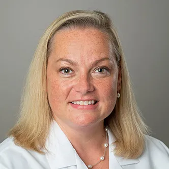 FNP Corbine has long blonde hair, is wearing a white medical coat and is smiling at the camera.
