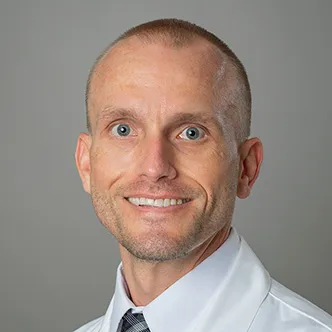 Dr. Davis has very short dark hair, is wearing a white dress shirt and tie under a white medical coat, and is smiling at the camera.