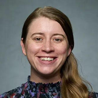 Dr. Dopp has long wavy brown hair worn in a side pony tail. She is wearing a dark purple and blue floral blouse and is smiling at the camera.