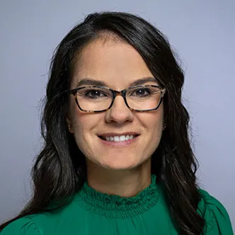 Dr. Hamersley has long wavy dark brown hair, is wearing glasses and a green blouse, and is smiling at the camera.