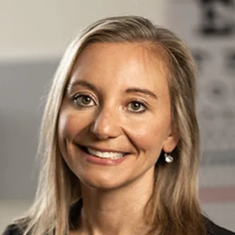NP Harris has long blonde hair, is wearing black medical scrubs while standing in an exam room. She is smiling at the camera.