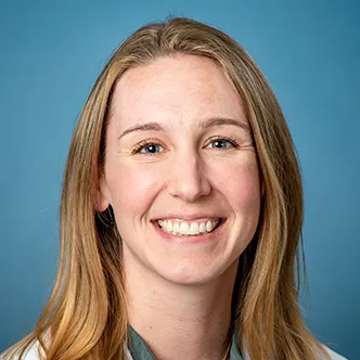 Jennifer Hayes has long reddish blonde hair, is wearing a blue blouse under a white medical coat, and is smiling at the camera.