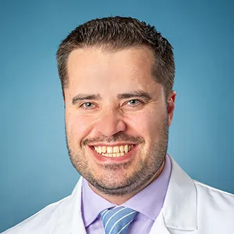 Carsten Kirby has short dark hair, a mustache and beard, is wearing a light purple shirt with blue tie under a white medical coat, and is smiling at the camera.