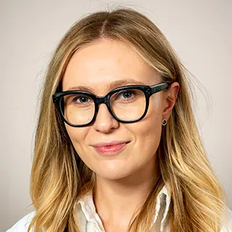 Dr. Kovalenko has long blonde hair, is wearing black rimmed glasses, a white medical coat, and is smiling at the camera.