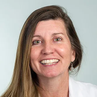 Dr. Lewis has long, straight, light brown hair, is wearing a white medical coat, and is smiling at the camera.