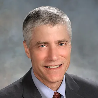 Dr. Netland has short gray hair, is wearing a gray suit with blue shirt and red tie, and is smiling at the camera.