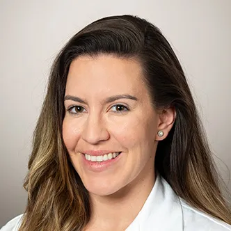 Dr. Olafson has long medium brown hair, is wearing a black blouse under a white medical coat, and is smiling at the camera.
