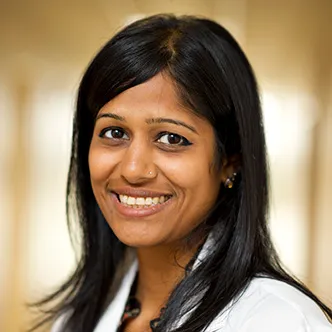 Dr. Patel has long, straight dark hair, is wearing a white medical coat, and is smiling at the camera.