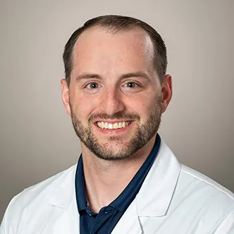 Dr. Petr has short dark hair and a beard, is wearing a dark shirt under a white medical coat, and is smiling at the camera.