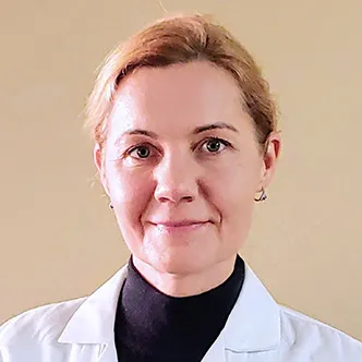 Anna Przybylska has blonde hair which is pulled back from her face, is wearing a dark turtleneck under a white medical coat and is smiling at the camera.