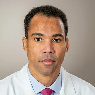 Dr. Tolentino has short dark hair, is wearing a blue shirt and red tie under a white medical coat and is looking at the camera.
