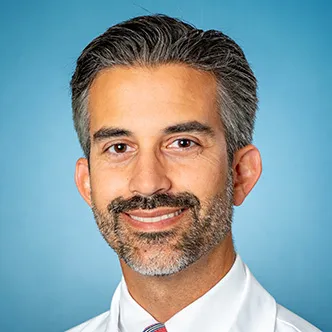 Dr. Volsky has short salt and pepper hair with a beard and mustache. He's wearing a white dress shirt and tie under a white medical coat and is smiling at the camera.