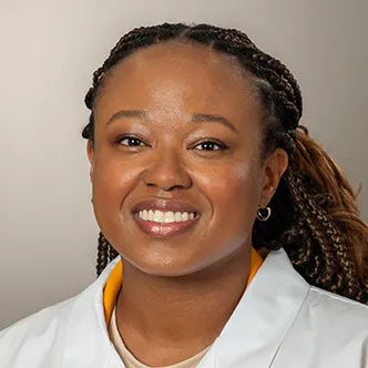 Ashley Walker has long, dark braids pulled back from her face, and is wearing a white medical coat while smiling at the camera.