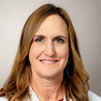 Janet Weir has long blonde hair parted on the side, is wearing a magenta blouse under a white medical coat, and is smiling at the camera.