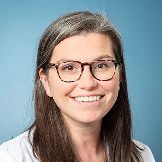 Dr.  Woll has long brown hair, is wearing glasses and a white medical coat and is smiling at the camera.