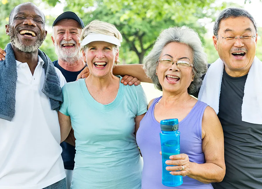 Five older adults of diverse ethnicity smile at the camera. They are in a park surrounded by green grass and trees, and are dressed for fitness activities.
