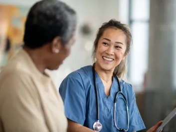 A female nurse sits with a senior patient wearing blue scrubs and has a tablet in her hands as she reviews some recent test results with her patient.