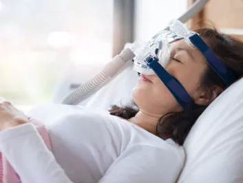 Senior woman using cpap machine to stop choking and snoring from obstructive sleep apnea with bokeh and morning light background.