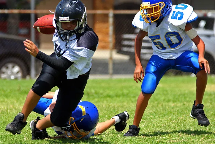 Three young people are wearing full gear including helmets, playing football on a grass field.