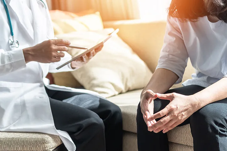 A doctor sits with a patient as they look at a tablet and talk in golden sunlight