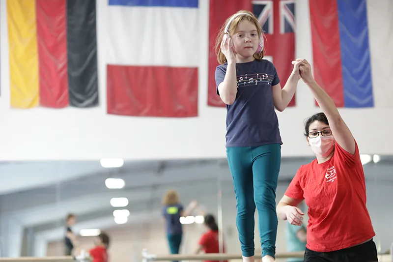 student walking on balance beam with headphones on holding hand of instructor. 