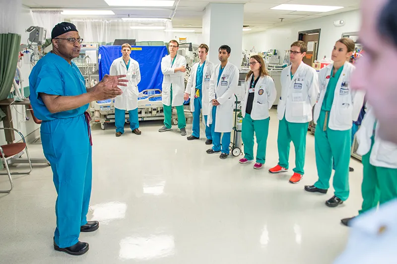 Clad in blue surgical scrubs, Dr Britt stands in a hospital hallway talking to large group of residents.