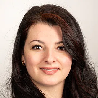 Dr. Jasim has long, dark, wavy hair, is wearing a white blouse under a white medical coat, and is smiling at the camera.