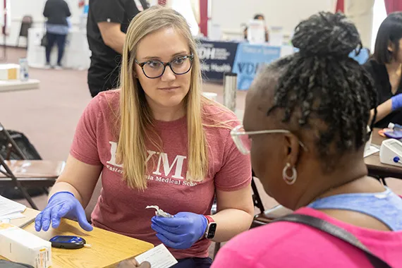 A medical student talks with a woman as she conducts a blood sugar test.
