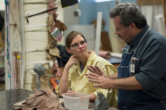 An instructor wearing an apron speaks to a student in a smock while standing over wet clay in an art studio.hile standing over