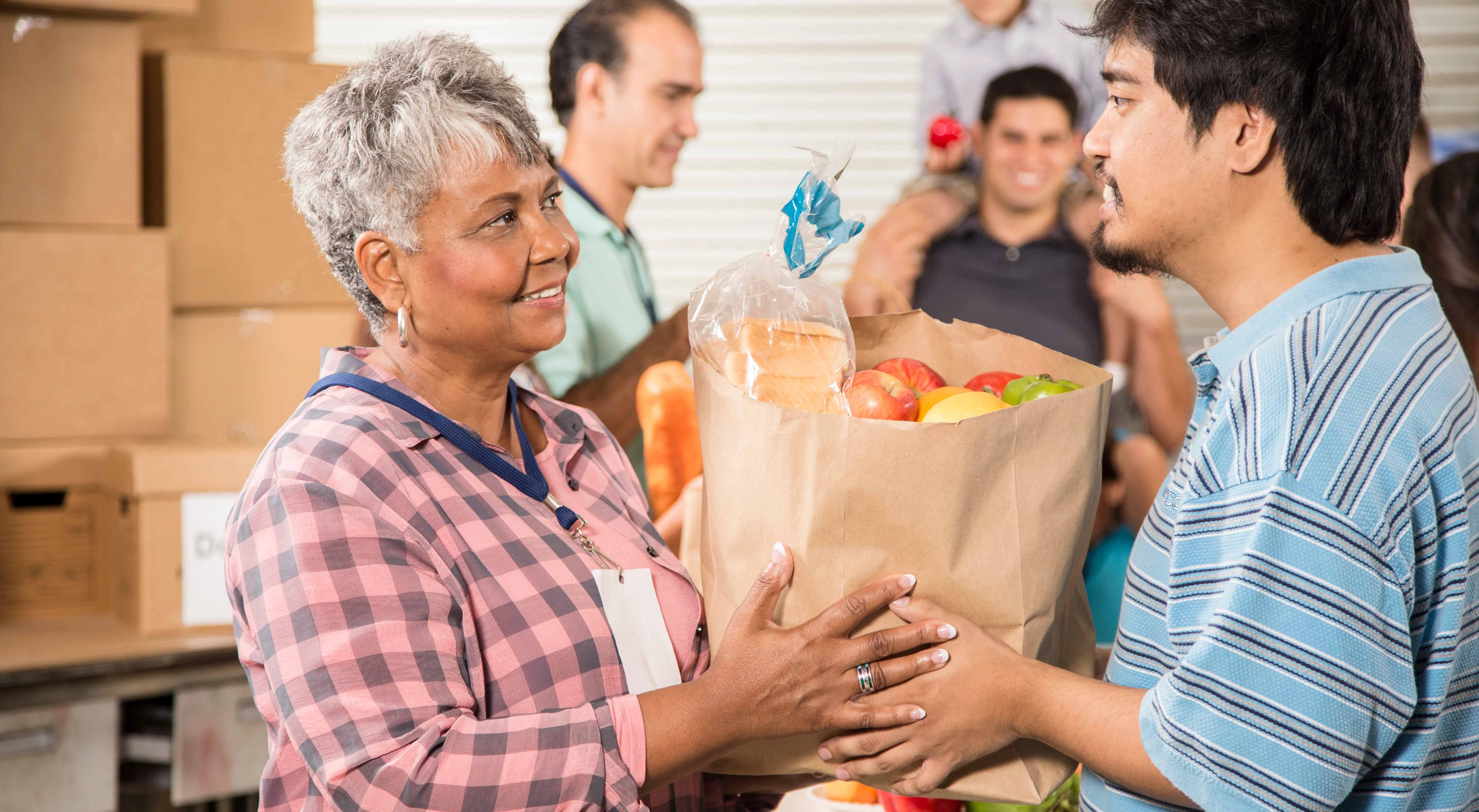 A food pantry employee hands a bag of food to another person, with several volunteer and clients in the background.