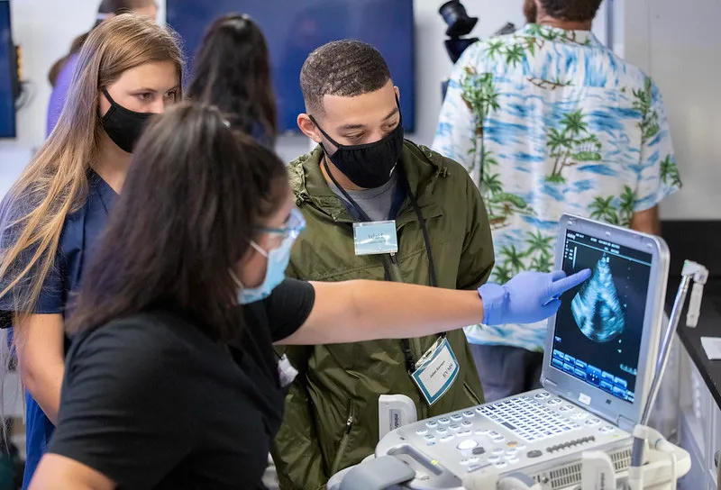 Three students wearing masks look at an ultrasound monitor