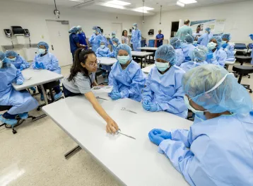 students and instructors wearing blue scrubs, gloves, and hair nets in classroom.