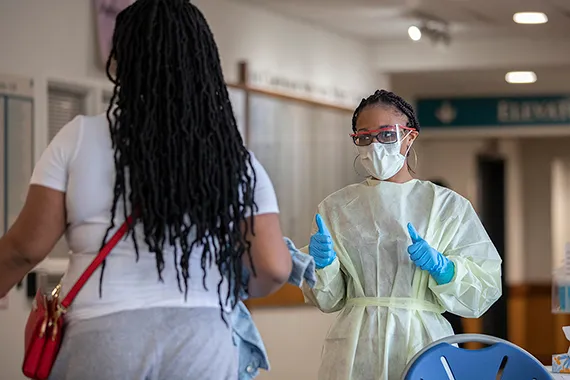 Screener wearing a mask and scrubs speaks to a person at an entrance