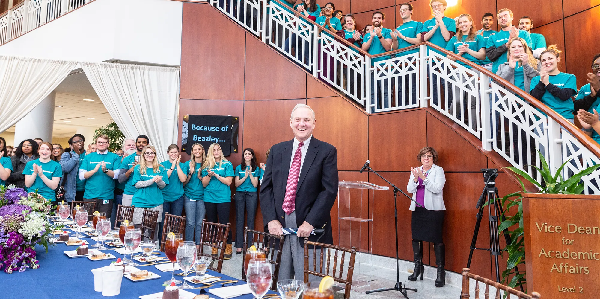 Judge Richard Bray stands in the center while dozens of students wearing teal shirts applaud, including many on the stairs behind him.