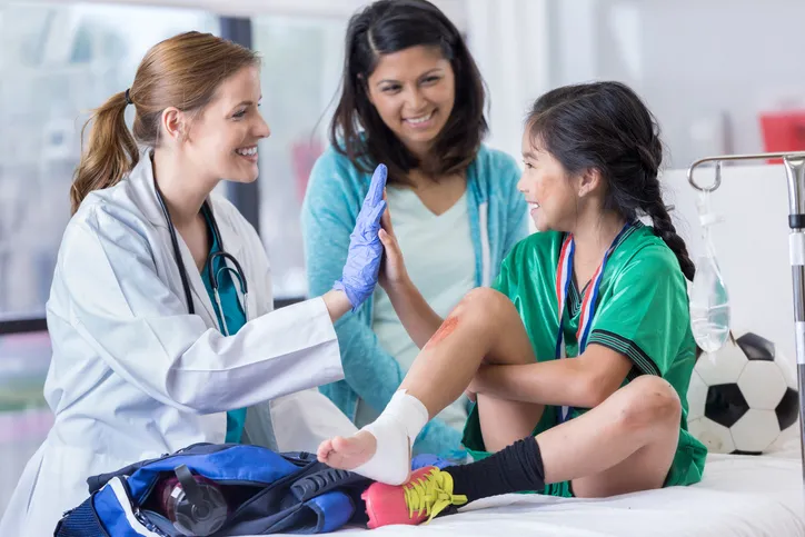 Doctor and an elementary age female patient high five one another. The girl has a sprained ankle. The girl's mom is smiling in the background.