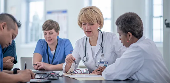A blonde female doctor in a white coat speaks to another female doctor, also in a white coat, both seated at a table