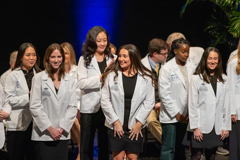 Students wearing white coats smile on stage during the ceremony