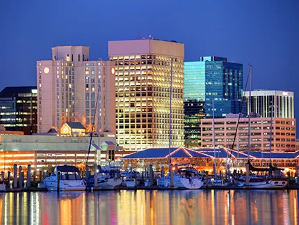 A luminous city skyline lies next to a river with boats against a dark blue evening sky