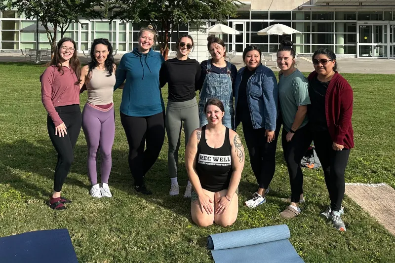 a group of individuals stand smiling together with yoga mats in front of Lester Hall.
