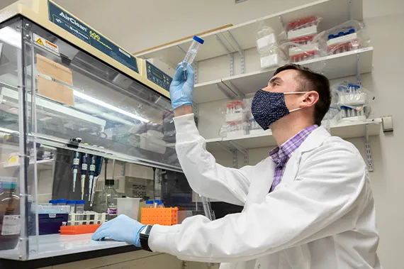 A male student wearing a PPE mask, lab coat and gloves works in a research lab.