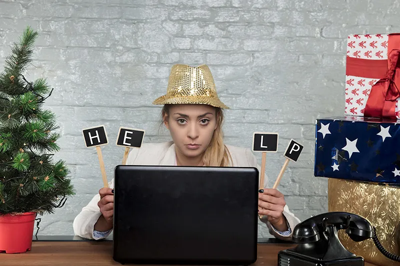 A woman is wearing a festive holiday hat surrounded by wrapped gifts and a green tree. She is holding a sign that reads HELP.