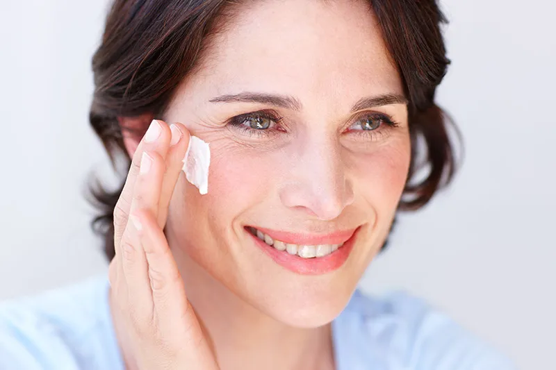 A mature woman with wavy brown hair smiles as she applies moisturizer to her face.